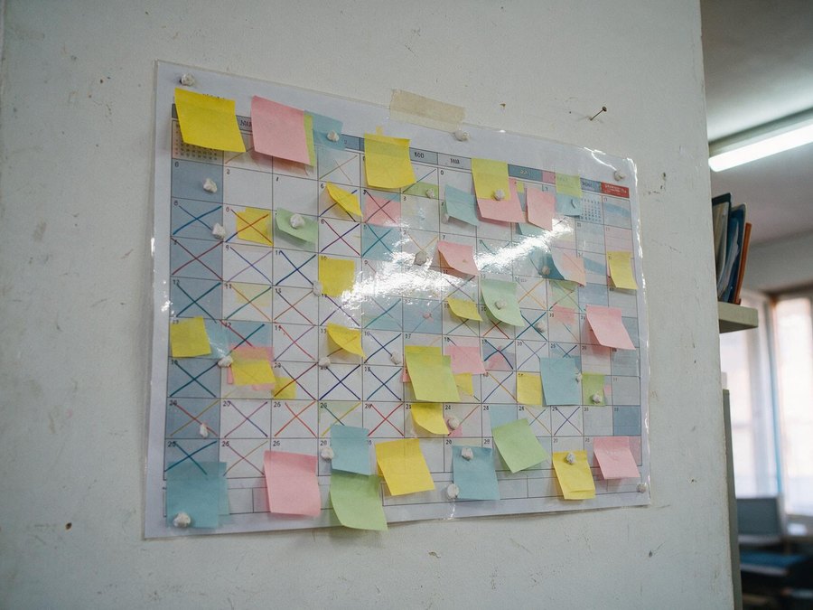 Un planificador mensual pegado a la pared de una oficina, cubierto de posits de colores y aspas de rotulador